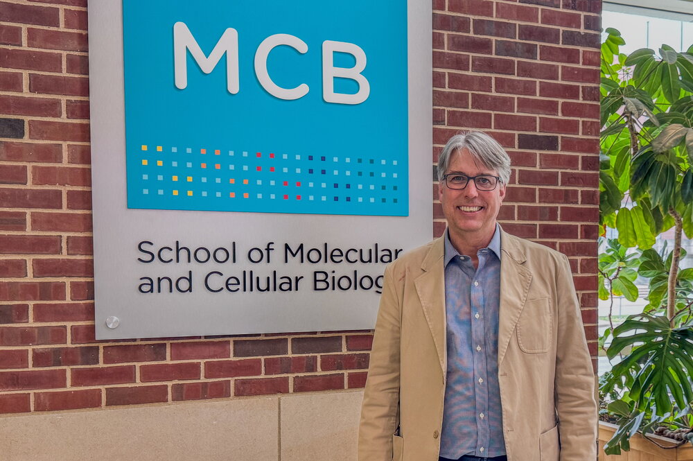 Man in blue shirt and beige jacket stands in atrium in front of sign that says MCB, School of Molecular & Cellular Biology